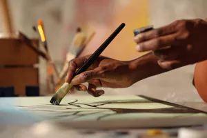 Close-up of hands painting on canvas. One hand holds a brush applying black paint, and the other holds a small jar. Brushes are in the blurred background.